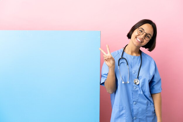 Mujer joven cirujano de raza mixta con una gran pancarta sobre fondo aislado sonriendo y mostrando el signo de la victoria