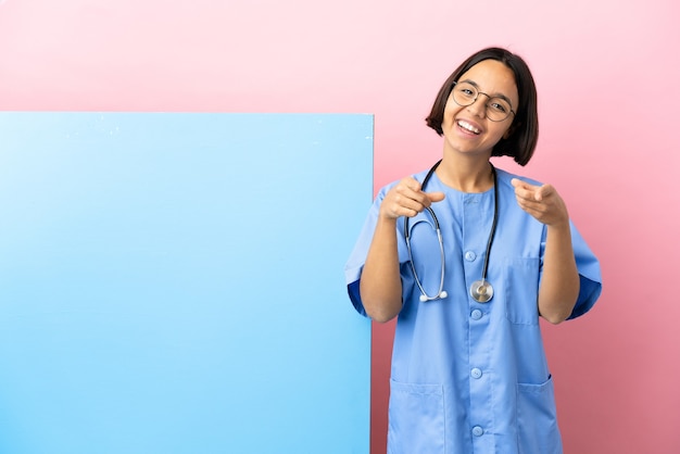 Mujer joven cirujano de raza mixta con una gran pancarta sobre pared aislada sorprendido y apuntando hacia el frente