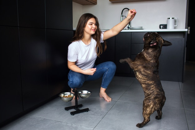 Foto mujer joven en la cocina durante la cuarentena. chica traning bulldog francés con comida para perros y jugando con mascotas. perro de piel oscura saltar.