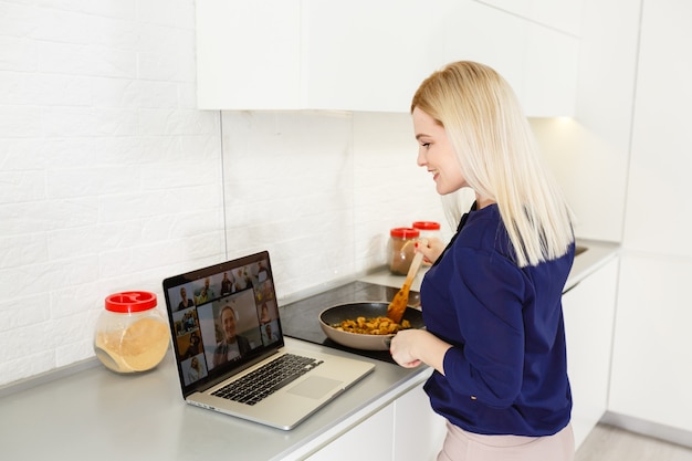 Mujer joven cocina en frente portátil abierto y videochating en la cocina