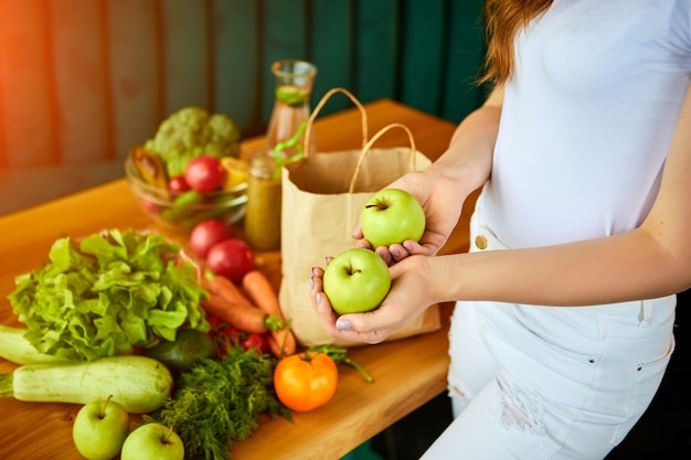 Foto mujer joven en la cocina sacando manzana de la bolsa de papel de compras con frutas y verduras.