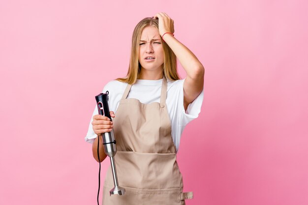 Mujer joven cocinera rusa sosteniendo una batidora eléctrica aislada cansado y con mucho sueño manteniendo la mano en la cabeza