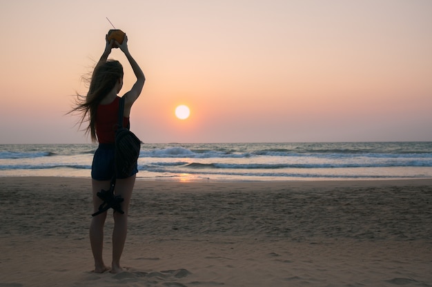 Mujer joven con un coco mira la puesta de sol