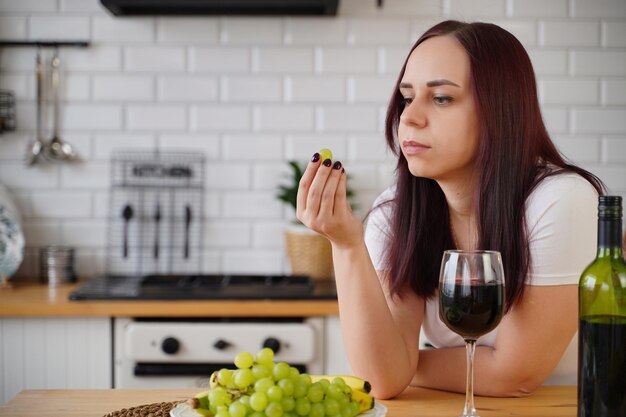 Mujer joven come uva verde en la cocina Hembra adulta descansando con frutas y alcohol en su fin de semana