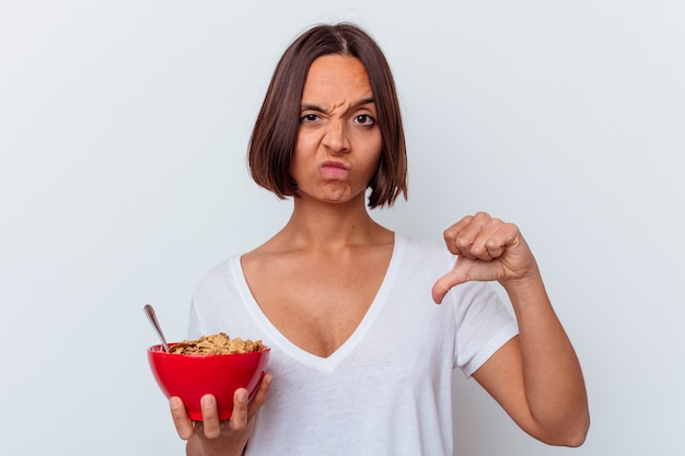 Mujer joven comiendo cereales aislado en la pared blanca mostrando un gesto de aversión, pulgares hacia abajo