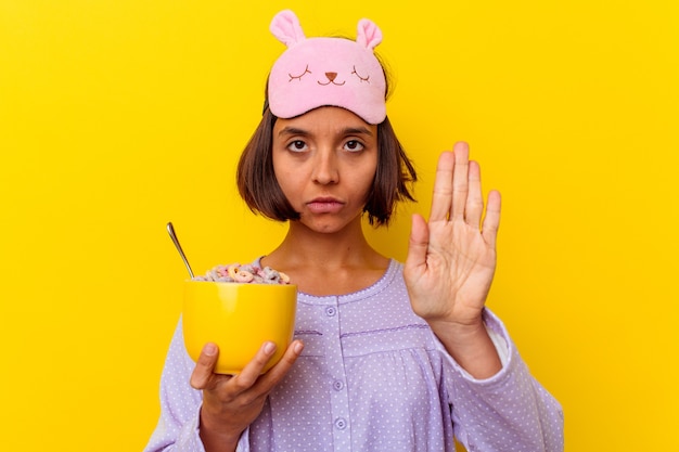 Mujer joven comiendo cereales vistiendo un pijama aislado en la pared amarilla de pie con la mano extendida mostrando la señal de stop, impidiéndole