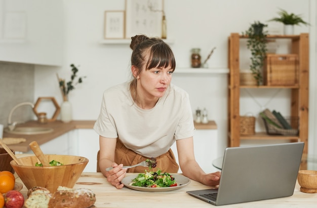 Mujer joven comiendo ensalada de verduras y usando la computadora portátil en la cocina