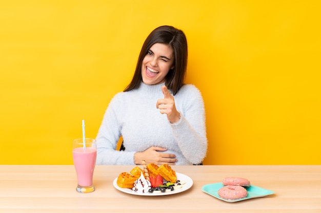 Mujer joven comiendo gofres y batido en una mesa sobre pared amarilla aislada le señala con el dedo