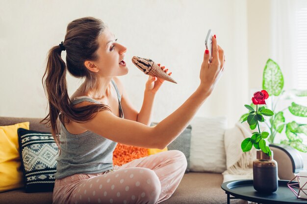 Mujer joven comiendo helado de chocolate en cono sentado en el sofá en casa y tomando selfie con smartphone.