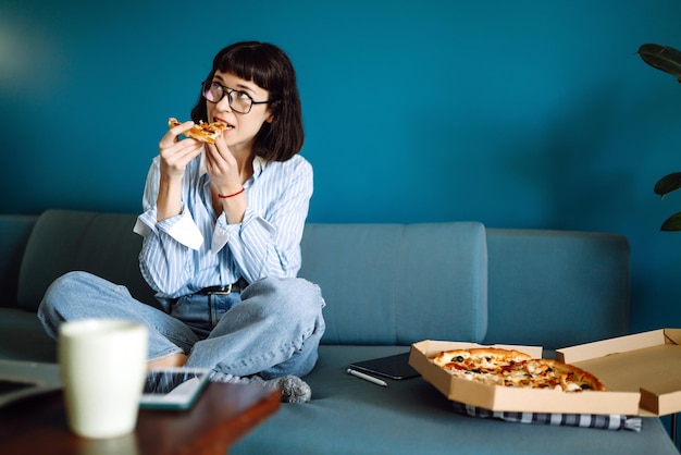 Mujer joven comiendo un pedazo de deliciosa pizza de la caja sentada en el sofá en casa Entrega de alimentos