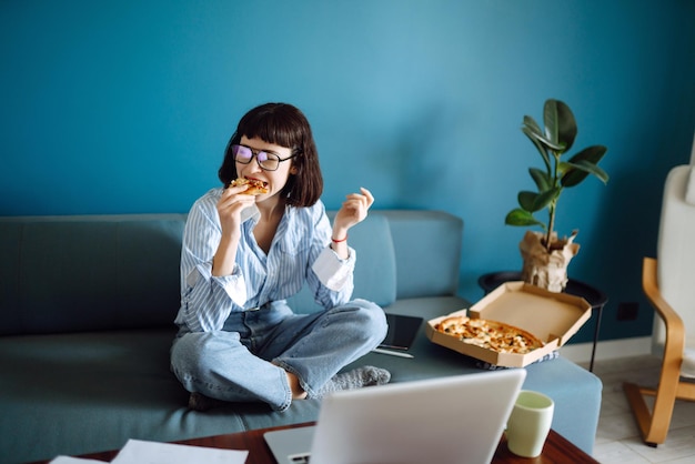 Mujer joven comiendo un pedazo de pizza deliciosa de la caja, sentada en el sofá en casa. Entrega de comida.