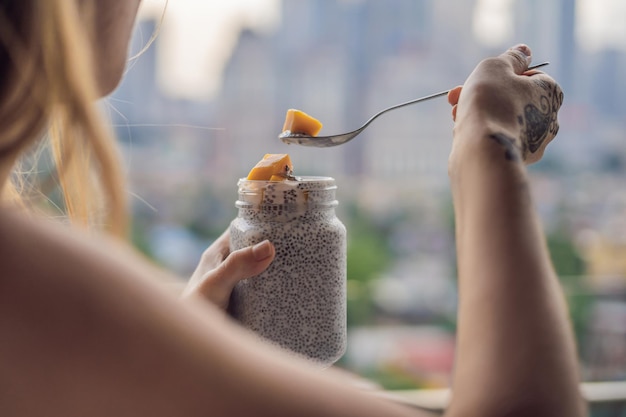 Mujer joven comiendo pudín de chía en su balcón con vistas a la gran ciudad