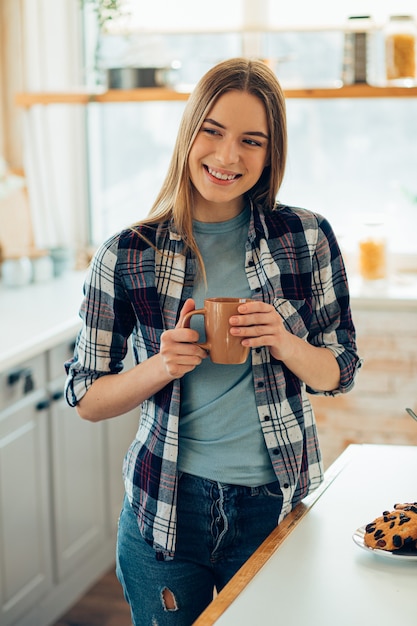 Foto mujer joven complacida de pie en casa con una taza en sus manos