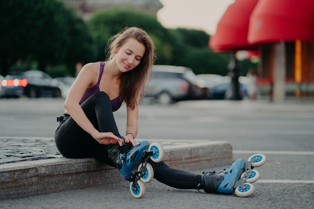 Mujer joven complacida se pone en patines para andar en rollos en un lugar urbano tiene ejercicio regular para deportes peligrosos ajusta cordones