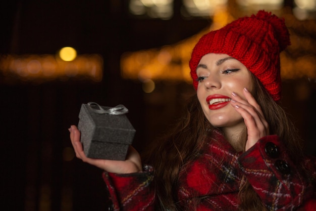 Mujer joven complacida con sombrero rojo recibiendo un regalo en una caja en la feria de Navidad por la noche. Espacio para texto