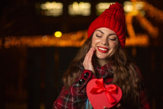 Mujer joven complacida viste gorra roja con caja de regalo con un lazo en la calle por la noche