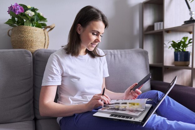 Mujer joven compra productos, cosméticos, ropa en línea. En manos del teléfono inteligente portátil con catálogo de papel, la mujer realiza compras en casa, sentada en el sofá