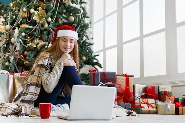 Mujer joven de compras en línea en la computadora portátil en el interior de la Navidad. Chica sentada bajo abeto decorado entre un montón de regalos envueltos. Preparándose para Navidad, pasando por las rebajas de invierno, copie el espacio