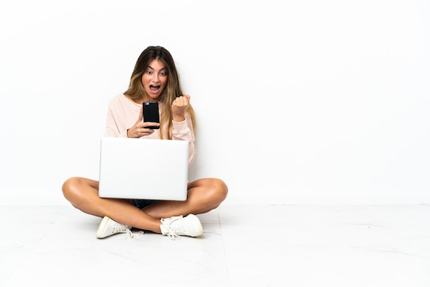 Mujer joven con una computadora portátil sentada en el suelo aislado en la pared blanca sorprendida y enviando un mensaje