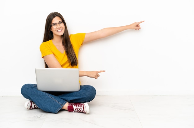 Mujer joven con una computadora portátil sentada en el suelo apuntando con el dedo hacia un lado y presentando un producto