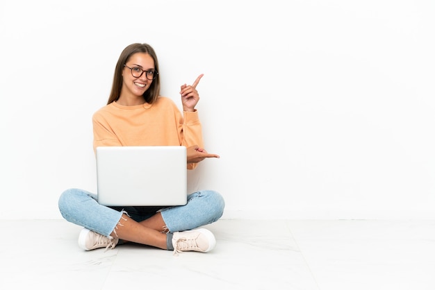 Mujer joven con una computadora portátil sentada en el suelo apuntando con el dedo hacia un lado y presentando un producto