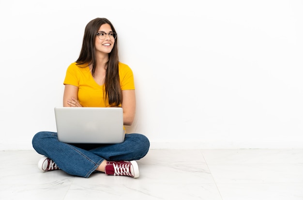 Mujer joven con una computadora portátil sentada en el suelo con los brazos cruzados y feliz