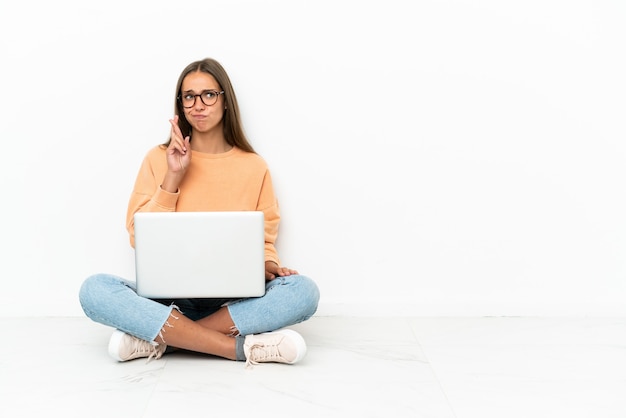 Mujer joven con una computadora portátil sentada en el suelo con los dedos cruzados y deseando lo mejor