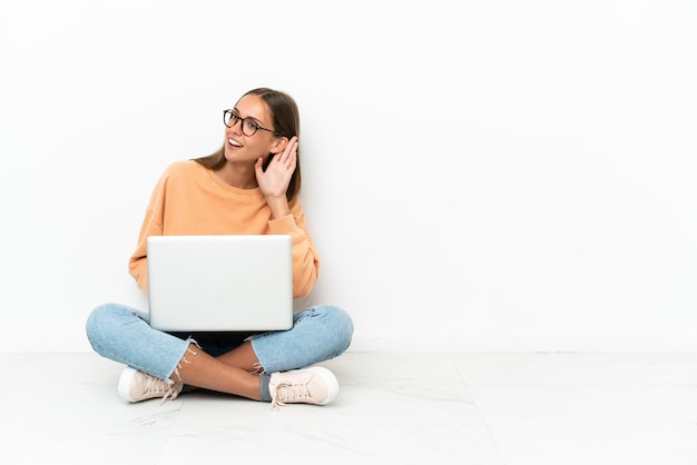 Foto mujer joven con una computadora portátil sentada en el suelo escuchando algo poniendo la mano en la oreja