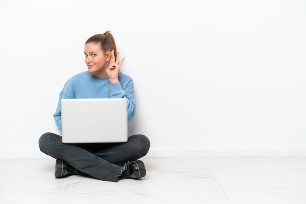 Mujer joven con una computadora portátil sentada en el suelo escuchando algo poniendo la mano en la oreja