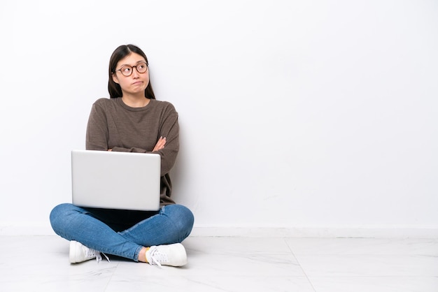 Mujer joven con una computadora portátil sentada en el suelo haciendo gestos de duda mientras levanta los hombros