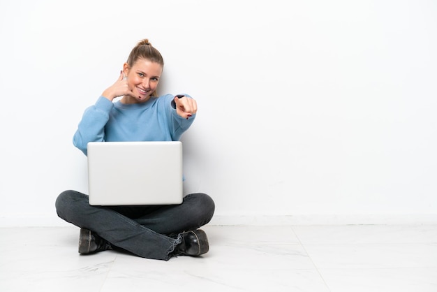 Mujer joven con una computadora portátil sentada en el suelo haciendo gestos de teléfono y apuntando hacia el frente