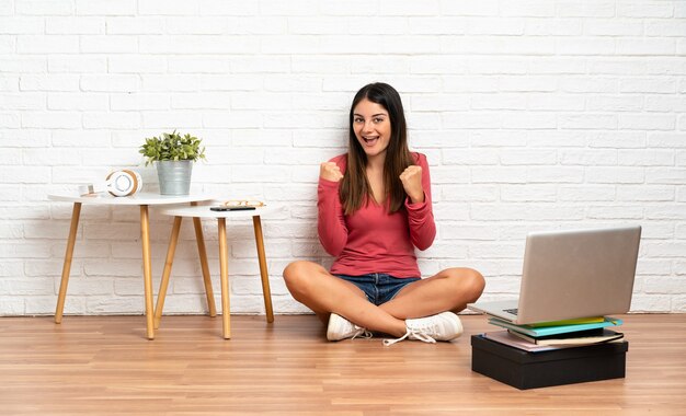 Mujer joven con una computadora portátil sentada en el suelo en el interior celebrando una victoria en la posición ganadora