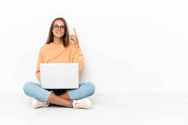 Mujer joven con una computadora portátil sentada en el suelo mostrando y levantando un dedo en señal de lo mejor