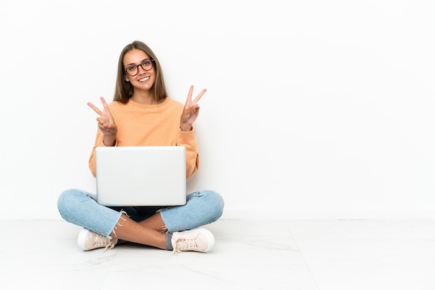 Mujer joven con una computadora portátil sentada en el suelo mostrando el signo de la victoria con ambas manos