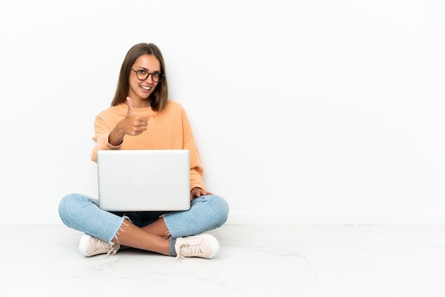 Mujer joven con una computadora portátil sentada en el suelo con los pulgares hacia arriba porque ha sucedido algo bueno