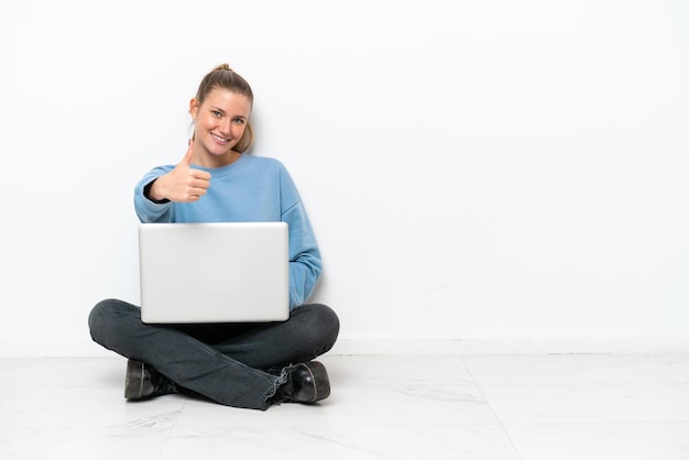 Mujer joven con una computadora portátil sentada en el suelo con los pulgares hacia arriba porque ha sucedido algo bueno