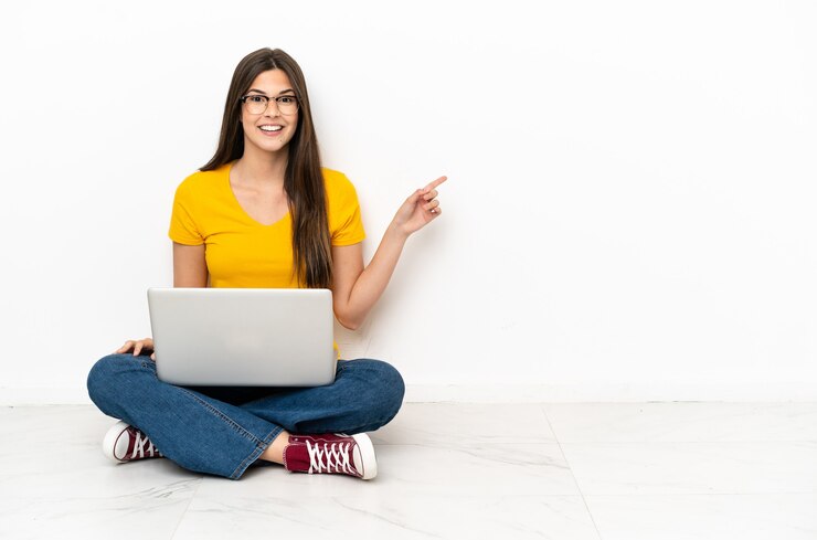 Foto mujer joven con una computadora portátil sentada en el suelo señalando con el dedo hacia el lado