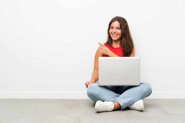 Mujer joven con una computadora portátil sentada en el suelo señalando con el dedo hacia el lado