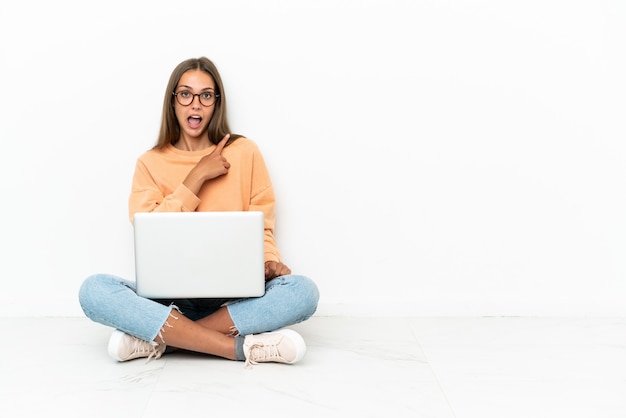 Mujer joven con una computadora portátil sentada en el suelo sorprendida y apuntando hacia el lado