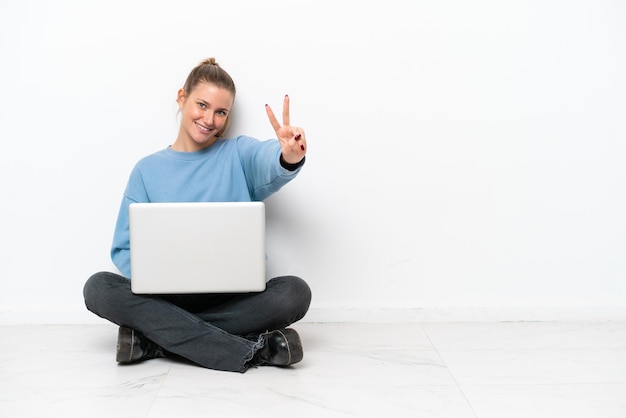 Mujer joven con una computadora portátil sentados en el suelo sonriendo y mostrando el signo de la victoria