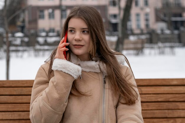 Mujer joven se comunica por gadget Chica morena con abrigo de piel de oveja se sienta en un banco en el parque de invierno y habla por teléfono inteligente
