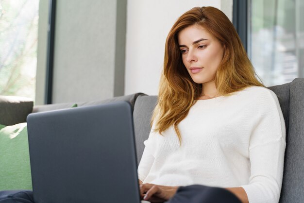 Mujer joven concentrada usando su computadora portátil moderna mientras está sentado en el sofá. Chica bonita de jengibre revisando mensajes mientras está sentado en casa. Concepto de vida doméstica
