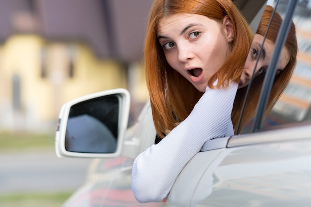 Mujer joven conduciendo un coche hacia atrás. Chica con expresión divertida en su rostro mientras hacía un daño en el guardabarros de un vehículo trasero.