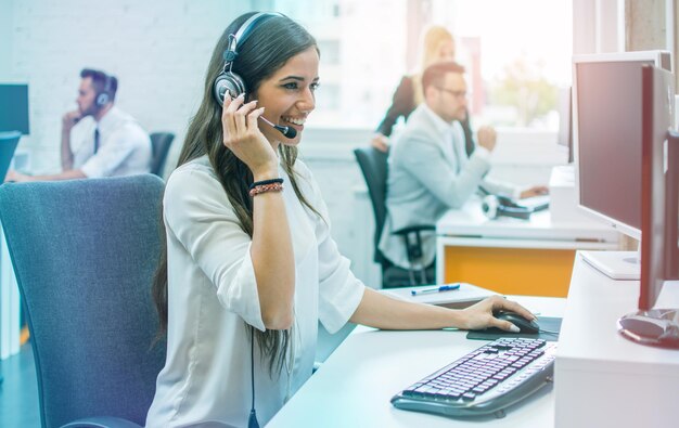 Mujer joven confiada con auriculares sentada en la oficina trabajando como operadora de centro de llamadas o servicio de apoyo con sus colegas