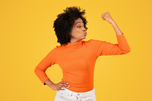 Mujer joven confiada con cabello afro flexionando el músculo del brazo en una postura fuerte con un naranja