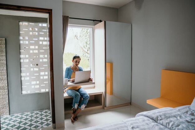 Foto mujer joven confiada en ropa casual elegante que trabaja en la computadora portátil mientras está sentado cerca de la ventana en casa