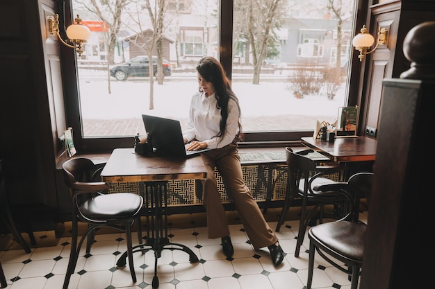 Mujer joven confiada en un traje de negocios trabaja en una computadora portátil mientras está sentada junto a la ventana en un café mujer empresaria