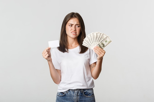Mujer joven confundida y disgustada mirando dinero y frunciendo el ceño, prefiere tarjeta de crédito.