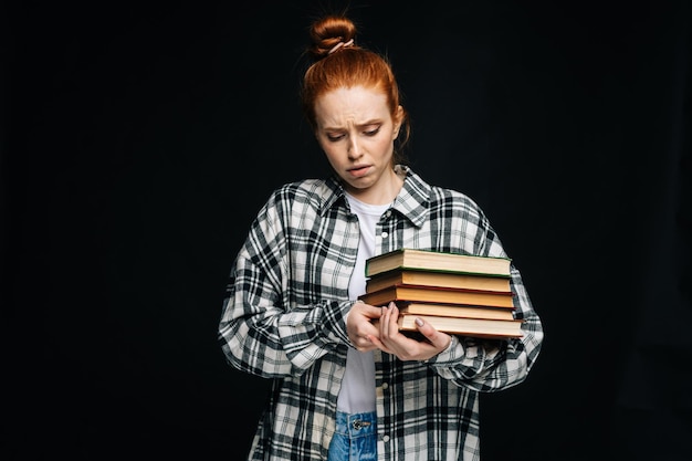 Mujer joven confundida estudiante universitaria sosteniendo un libro y llorando sobre un fondo negro aislado Bonita pelirroja modelo vistiendo ropa casual mostrando emocionalmente expresiones faciales