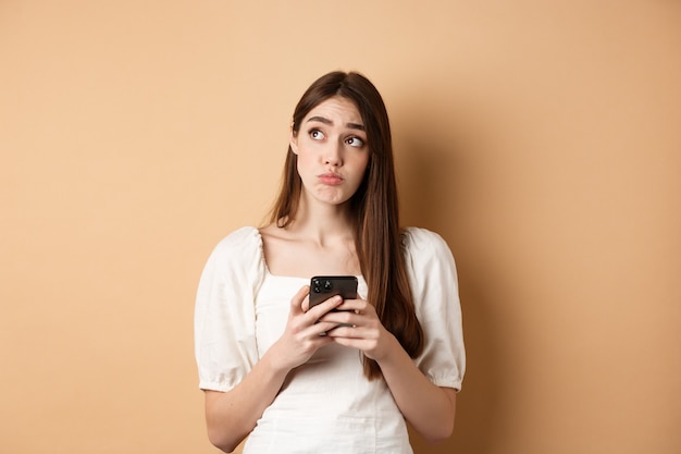 Mujer joven confundida pensando después de leer noticias en el teléfono móvil, mirando la esquina superior izquierda vacilante, de pie sobre fondo beige.